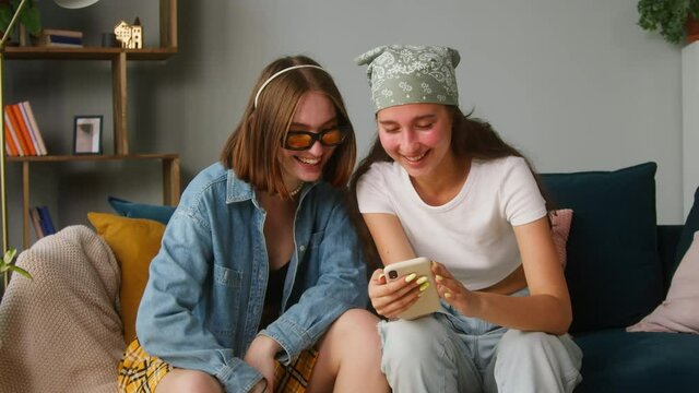 Young women sitting on sofa and watching funny videos using mobile phone. Two sisters watch stories in social networks. Female friends surfing the internet, reading news on smartphone and laughing.