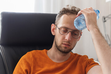 Working at home man suffering from heat and thirst cools down with water bottle at hot summer day - obrazy, fototapety, plakaty
