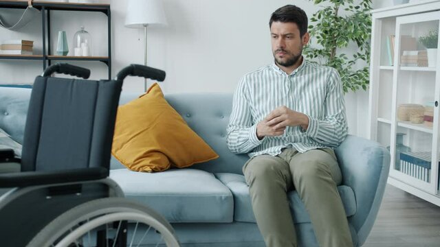 Slow motion portrait of miserable handicapped man sighing sitting on sofa with wheelchair in forefront. Depression and disabled people concept.