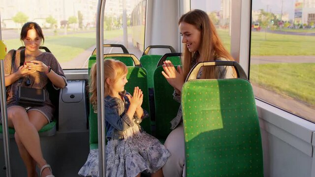 Family rides in public transport tram, mother with blonde girl sit together and playing game clapping hands, talking, laughing. Woman passenger with her daughter having fun in bus. Commuters. Train