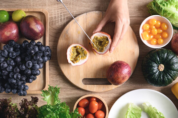 Hands of woman holding knife and cutting passion fruit on wooden board with fresh vegetables for...
