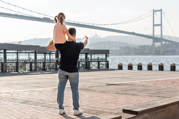 Little cute girl seat on father's shoulders in front of Bosphorus bridge (aka: 15 July Martyrs Bridge Turkish: 15 Temmuz Sehitler Koprusu) in Ortakoy, Istanbul, Turkey. Man with daughter.
