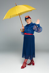 smiling elderly woman in blue dress and turban holding yellow umbrella and waving with hand on grey