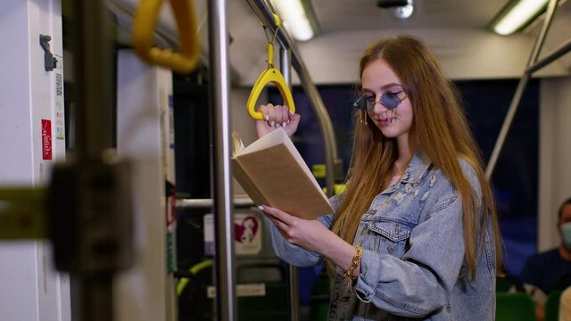 Portrait of attractive young adult woman stay at empty subway train and reading interesting book. Concept of people, literature and public transport. Girl passenger traveling at modern tram or bus