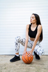 Fit young woman posing in front of white garage door with basketball ball 