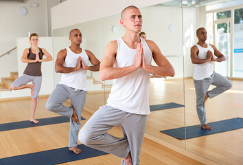 Obraz premium Concentrated man standing on one leg in Eka Pada Pranamasana pose during group yoga training in gym