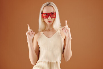 Cool young Caucasian woman in red striped goggles puckering lips and pointing fingers up against brown background
