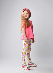 Red-haired kid girl in summer wear pink t-shirt, colorful pants and sandals stands sideways looking back over her shoulder. Side view. Full-growth