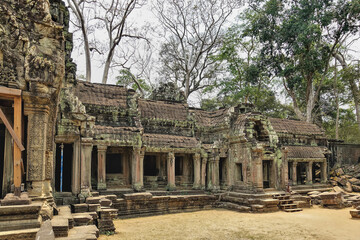 Obraz premium Ruins of a temple in the ancient city of Angkor. A dilapidated roof and galleries with columns. Bas-reliefs and carvings on weathered stones. Jungle trees against the sky. Cambodia