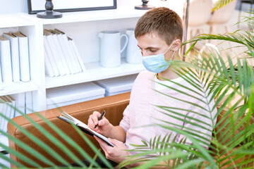 cute Caucasian man in a protective mask sits in a chair with a notebook in his hand and writes, remote work and social distance learning