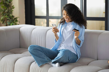 Happy young african american woman with curly hair uses credit card and mobile phone for online...