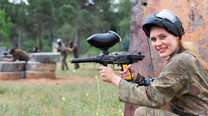 Young smiling woman paintball player in camouflage posing with gun outdoors