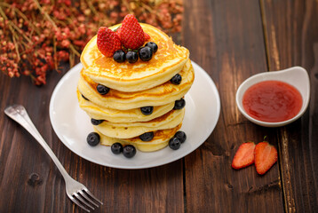 Sweet food. Stack of delicious pancakes with blueberries, Strawberry and Strawberry jam, fork rests in white plate on blur wooden background.