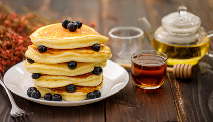 Sweet food. Stack of delicious pancakes with fork rests blueberries and honey, teapot in white plate on blur wooden background.