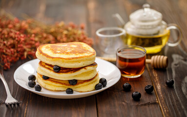 Sweet food. Stack of delicious pancakes with fork rests blueberries and honey, teapot in white plate on blur wooden background.