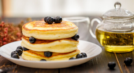 Sweet food. Stack of delicious pancakes with fork rests blueberries and honey, teapot in white plate on blur wooden background.