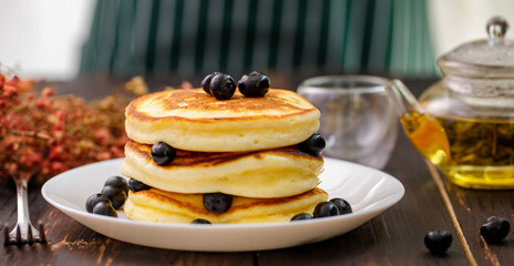 Sweet food. Stack of delicious pancakes with fork rests blueberries and honey, teapot in white plate on blur wooden background.