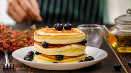 Close up hand of unrecognizable woman holding hand decorating Pancake by picking up blueberries and blur teapot background. Concept sweet food.