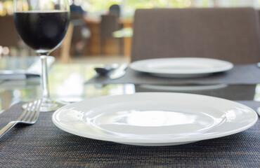 Selective focus and blurred background of tableware clean set (white plate or dish, fork and spoon) with glass of red wine prepared for food of lunch meal in luxury formal restaurant.