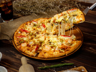 Selective focus and close up on a piece of double cheese homemade seafood pizza on spatula or scoop with blur background of wooden table, sackcloth, ingredients and fried potatoes. Food Concept.