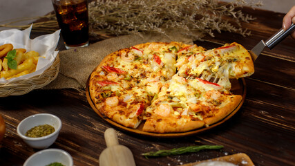 Selective focus and close up on a piece of double cheese homemade seafood pizza on spatula or scoop with blur background of wooden table, sackcloth, ingredients and fried potatoes. Food Concept.