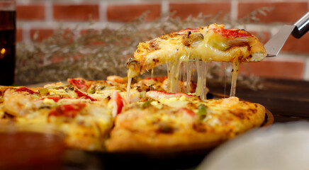Selective focus and close up on a piece of double cheese homemade seafood pizza on spatula or scoop with blur background of wooden table, cold beverage and brick wall. Food Concept.