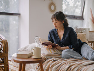 Young relaxed and happy Asian woman in casual outfit reading book and drinking tea while relaxing on sofa in cozy living room at home, enjoying free time on weekend at home