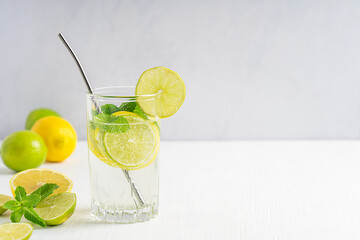 Refreshing homemade lemonade made of lemon and lime slices, sparkling water and green mint served in glass with metal straw on white wooden table against gray wall. Image with copy space, horizontal