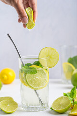 Glass of refreshing homemade lemonade made of lemon slices, sparkling water and mint served with metal straw on white wooden table at kitchen and hand on the top squeezing slice of lime. Vertical