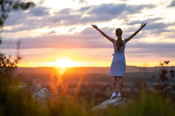 Fototapeta premium Dark silhouette of a young woman standing with raised up hands on a stone enjoying sunset view outdoors in summer.