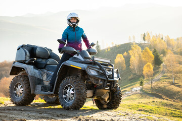 Fototapeta na wymiar Happy active female driver in protective helmet enjoying extreme riding on ATV quad motorbike in fall mountains at sunset.
