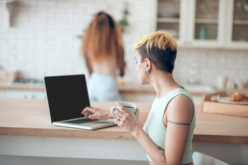 Woman drinking coffee at laptop in kitchen and girlfriend