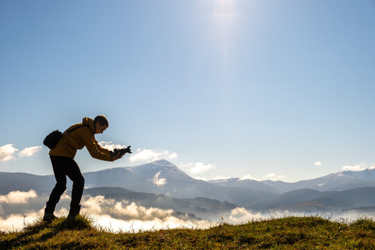 Silhouette of a backpacker photographer taking pictures of morning landscape in autumn mountains with digital camera.