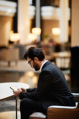 Side view portrait of bearded Middle-Eastern businessman using smartphone while waiting in hotel lounge