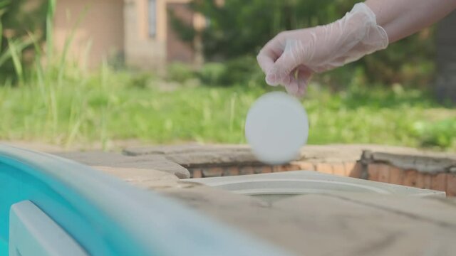 Disinfection and purification of water in swimming pool. Hand in protective glove throwing white chlorine tablet in pool skimmer closeup.