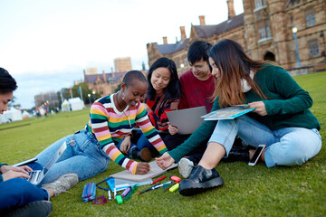 Group of young university students hanging out sitting on grass studying and using devices