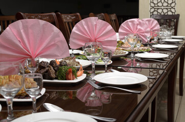 Large brown festive banquet table is decorated with pink napkins and served with white plates, spoons, forks, wine glasses, festive salads and snacks from vegetables and meat are on the table