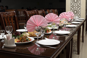 Large brown festive banquet table is decorated with pink napkins and served with white plates, spoons, forks, wine glasses, festive salads and snacks from vegetables and meat are on the table