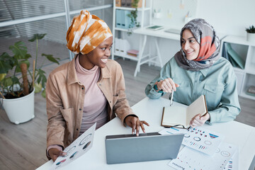 High angle portrait of two ethnic young women wearing headscarf while enjoying work together in...