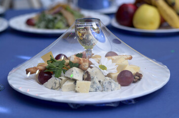 Glass with a different snack of cheese, cheese, grapes, walnuts, bread sticks on a plate, covered with a transparent film, on a blue banquet table