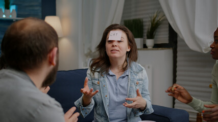 Multiethnic friends sitting on sofa late at night playing guess who game attaching sticky notes on forehead. Group of multiracial people spending time together having fun during entertainment wekeend