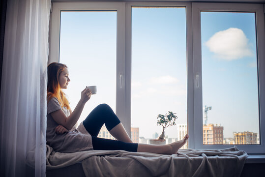 Silhouette of young slender girl sitting on the windowsill at home, side view, copy space. Outside the window blue sky and tall city buildings. Red-haired woman holding cup, looking at the metropolis.