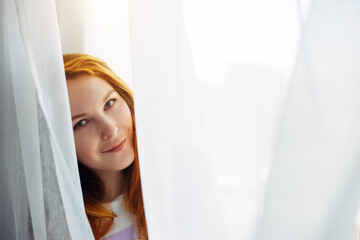 Cute young red-haired girl looks out from the white curtain, close-up, copy space. Portrait of beautiful smiling woman with perfect skin on a light blurry background.