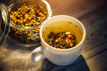 Beautiful blended tea made of herbs, berries and nuts in white mug and transparent glass bowl on vintage wooden background, close-up. Brewing tea in the evening light. Healthy lifestyle, detox.