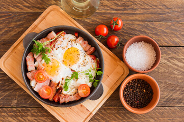 Homemade fried eggs with bacon and tomatoes in a frying pan on wooden background. Classic breakfast. Top view.