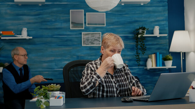 Older Professional Mature Business Woman Using Laptop Sitting At Workplace Desk Drinking Coffee. Retired Remote Executive Manager Working From Home Typing On Pc, While Husband Watching Tv In