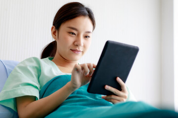 An asian young pretty female patient smiling with happiness and touching screen of tablet for online chat or communicate while lying on bed in hospital. Technology and Medical Concept.