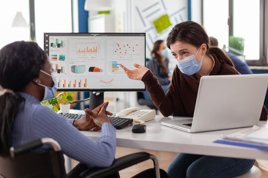 Freelancers wearing protective face masks working on computer in business office during global pandemic, disabled african woman sitting in wheelchair . Team discussing in new normal financial office.