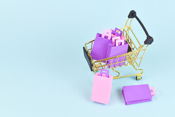 Small golden shopping cart filled with pink and purple paper shopping bags on side of blue...