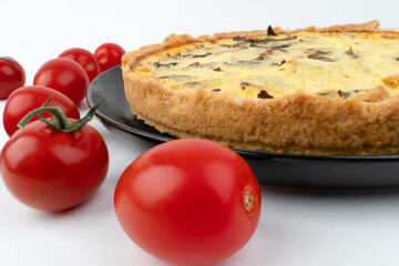 Closeup wide studio shot of freshly baked yellow French salty cake, or quiche, with mushrooms, red cherry tomatoes, on a black design plate, isolated on white background